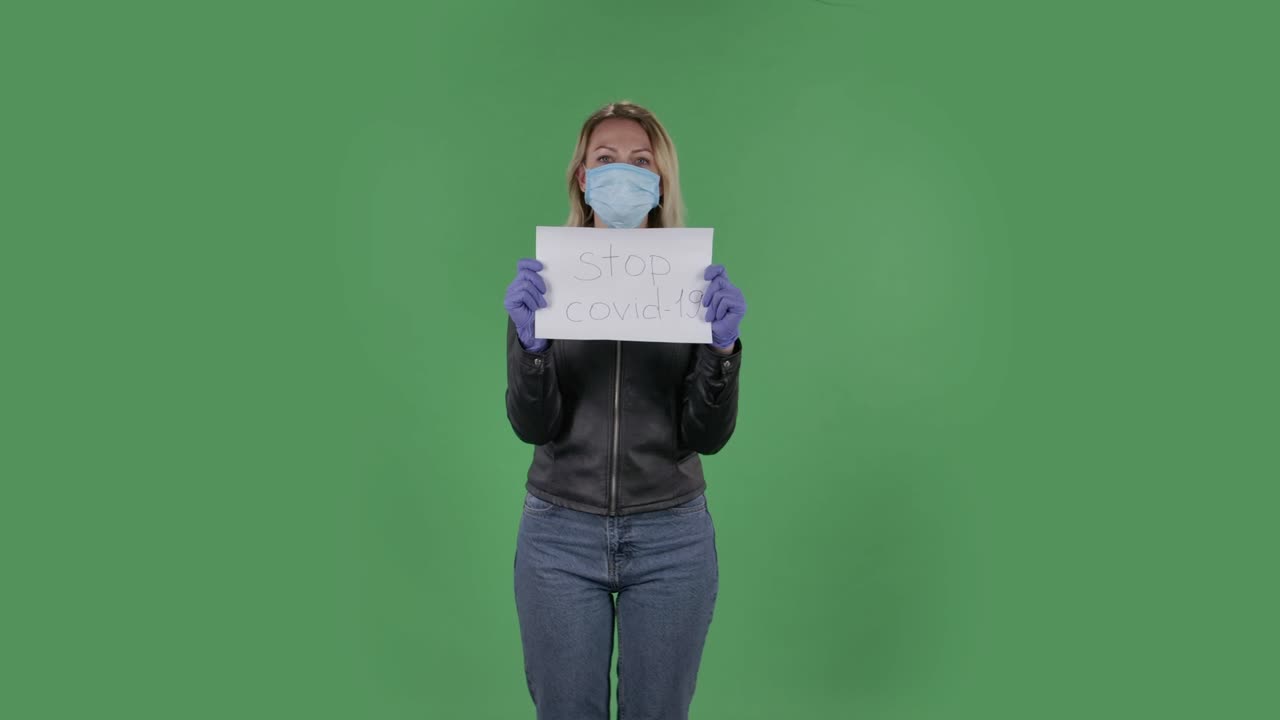 의료 마스크를 입은 아름다운 젊은 여성의 초상화는 카메라를 바라보고 코로나19를 멈추는 포스터를 들고 있습니다. 검은색 재과 청바지를 입은 느슨한 머리카락을 가진 금발은 스튜디오에서 녹색 화면에 있습니다. 건강 보호 코로나 바이러스 개념