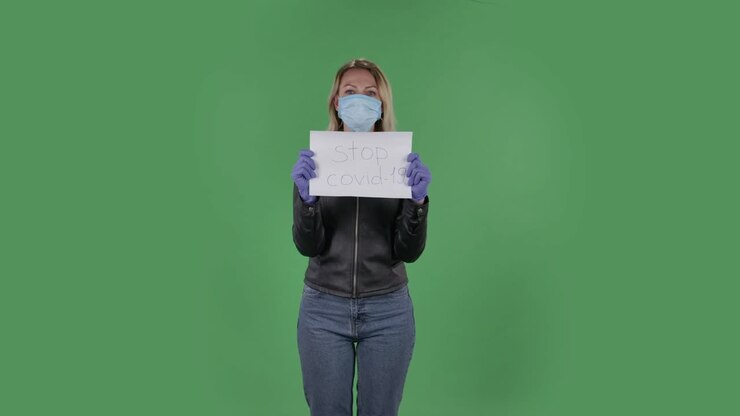 Portrait of beautiful young woman in medical mask is looking at camera and holding a poster Stop Kovid-19.Blonde with loose hair in a black jacket and jeans on a green screen in the studio. Health Protection Corona Virus Concept