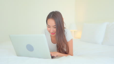 close-up of an attractive woman working on her laptop as she lays on a big comfortable bed