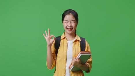 asian woman student with a backpack and some books smiling and showing okay gesture while standing in the green screen background studio