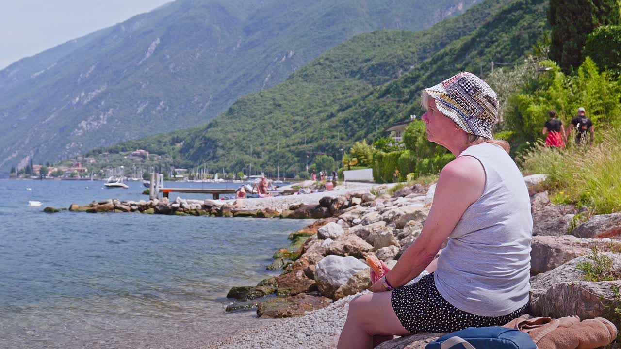 Woman Eats Picnic Sandwich Sitting on sunny rocky shores Lake Garda