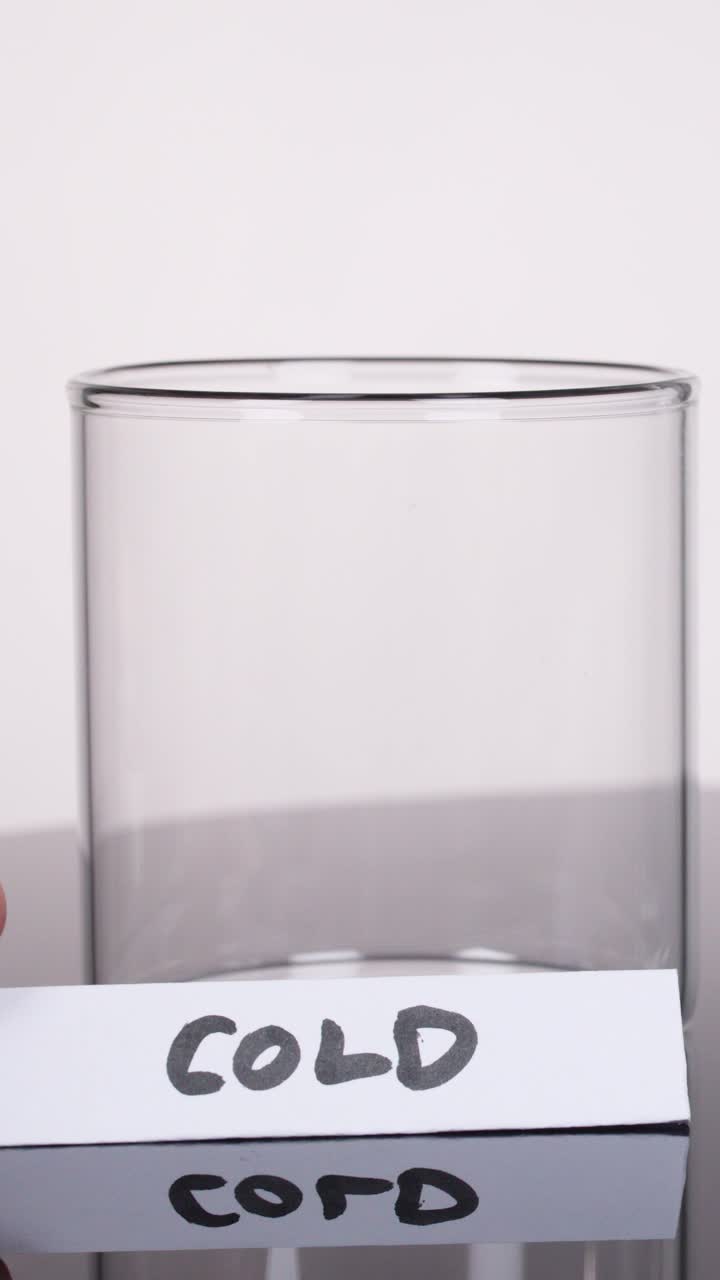 Una mano posiziona l'etichetta 'FREDDO' accanto a un bicchiere vuoto su una superficie riflettente, sotto l'illuminazione da studio, inquadratura statica.