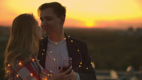 Man-and-young-pretty-woman-sitting-on-couch-holding-wine-glasses-and-kissing-on-rooftop-terrace-at-sunset