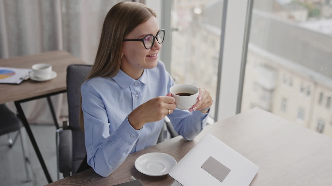 A little break from work to have a cup of coffee. Business lady drinks coffee sitting in the office enjoying view from panoramic window.