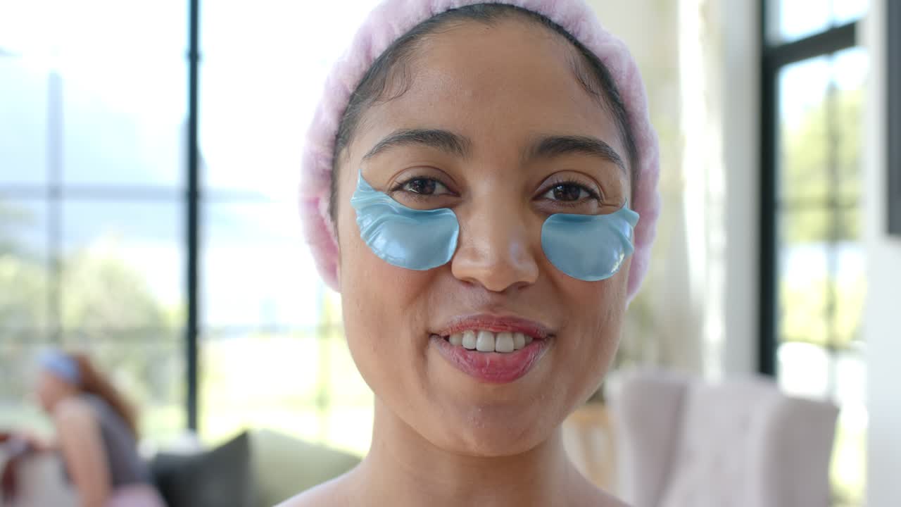 Amigas afroamericanas en un spa casero, una amiga sonriendo y hablando a la cámara con parches azules y una diadema rosa.