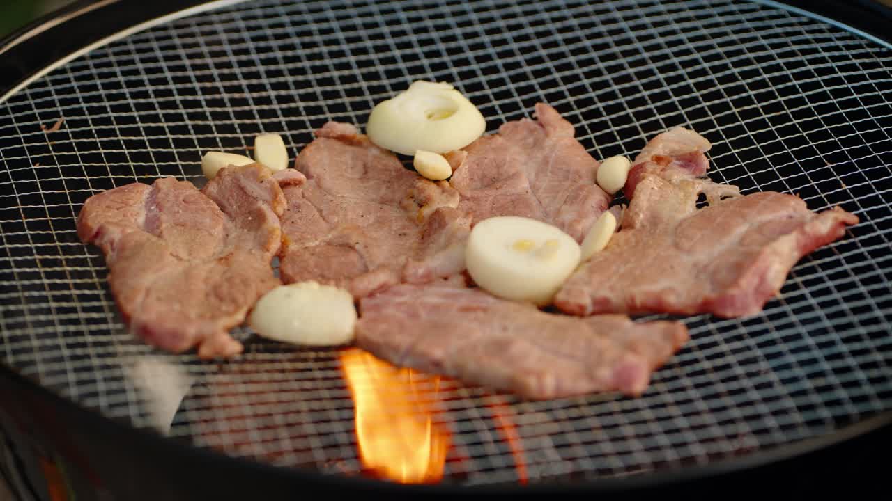 Close-up shot of a person using tongs to add fresh garlic cloves to delicious thin-sliced pork cooking on a hot wire mesh grill over a flaming charcoal barbecue