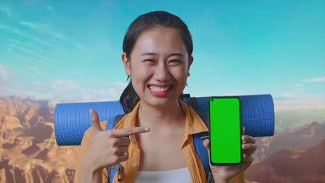 Close Up Of Asian Female Hiker With Mountaineering Backpack Smiling And Pointing To Mock Up Green Screen Smartphone While Traveling At The Top Of Mountain