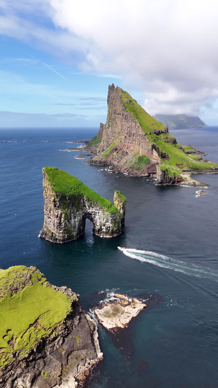 Cinematic aerial view of Drangarnir sea stacks rising dramatically from the Atlantic Ocean near Vágar, Faroe Islands, showcasing rugged cliffs, lush green slopes, and misty Nordic seascape