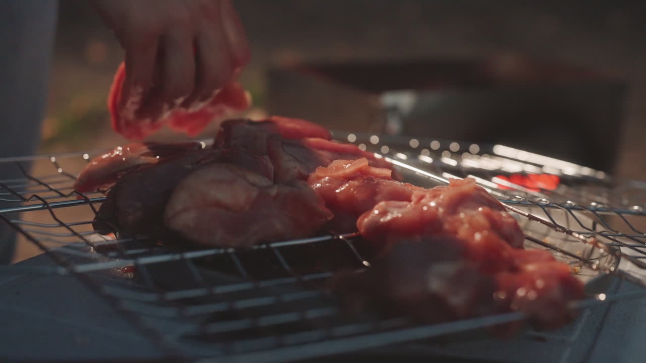 Panggangan berkemah saat senja dengan ayam mentah, tangan meletakkan potongan yang sudah dimarinasi di atas kawat panggangan, pijaran arang dan bokeh lembut, masakan luar ruangan yang intim, suasana pedesaan, persiapan makanan yang terasa, tampilan dekat gerak lambat.