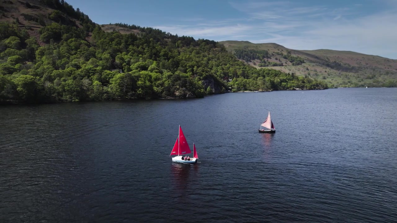 Barca a vela con vela rossa nel Lake District inglese sul lago Ullswater, Cumbria, Inghilterra. Una piccola isola in primo piano. Tempo soleggiato e luminoso.