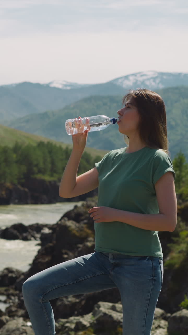 Tired woman drinks water from bottle and looks around on rocky mountain river bank during hike. Avoid dehydration and heat stroke. Health care