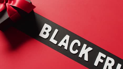 Black-friday-text-in-white-on-black-stripe-and-black-gift-box-with-red-ribbon,-on-red-background