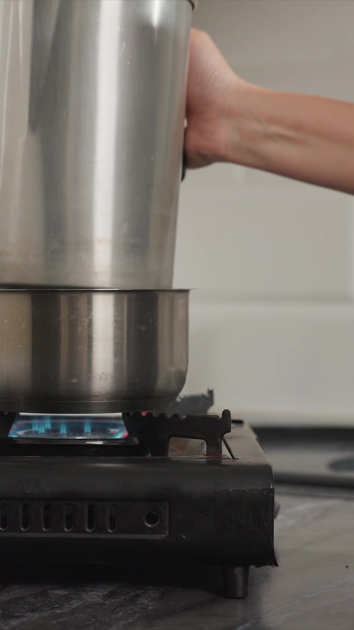 Close up of stainless steel pot heating on gas stove with blue flame showing steam rising from boiling water. hand places another metal container over heated bottom one to create double