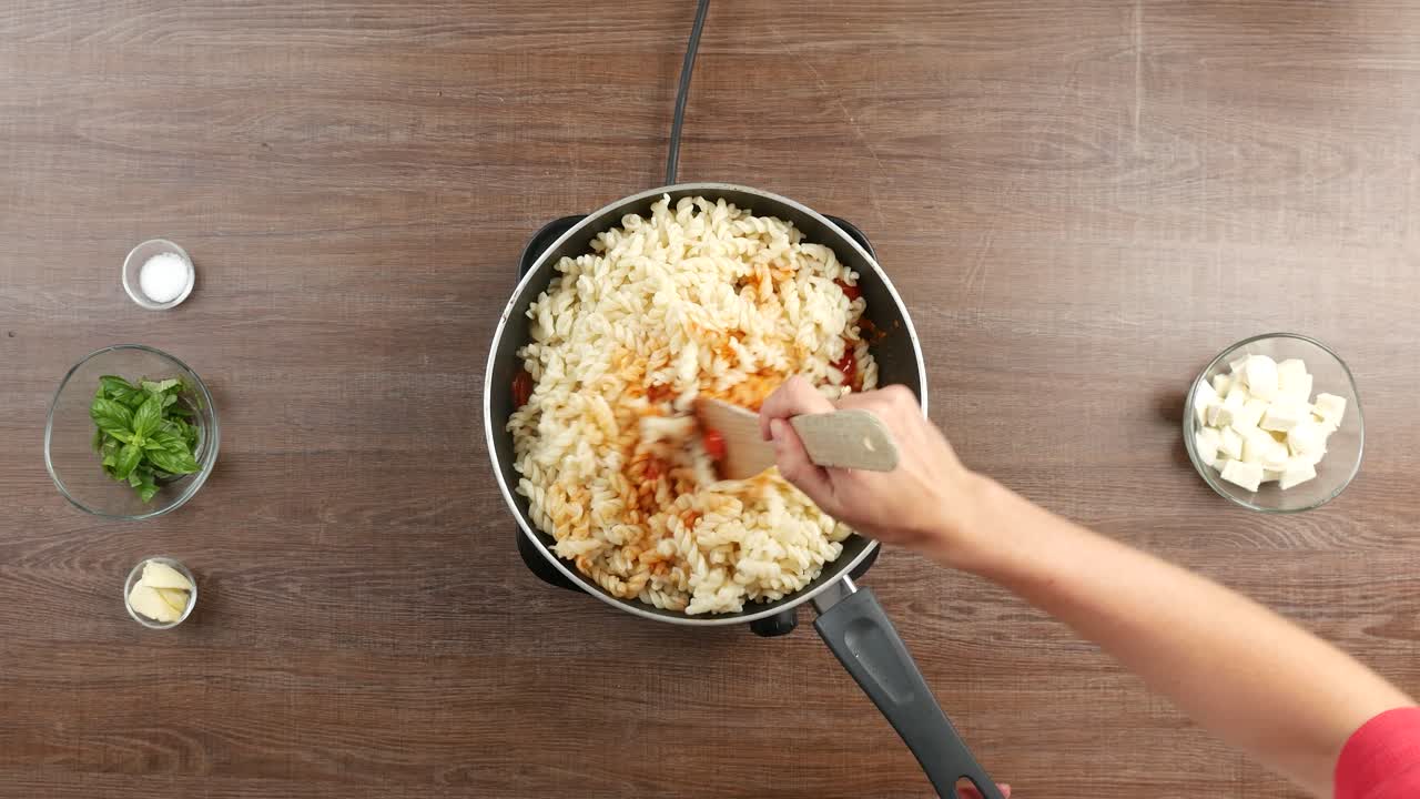 toma de arriba hacia abajo de una combinación de pasta fusilli con salsa de tomate en una sartén grande, usando una espátula de madera.