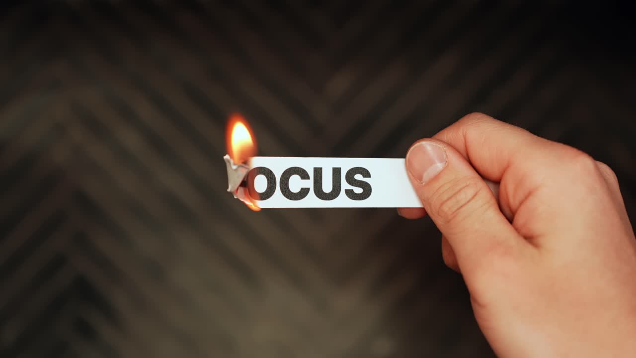 Close-up hand holds a paper label reading “FOCUS” as it catches fire, symbolizing burnout, distraction, and lost productivity on a dark background