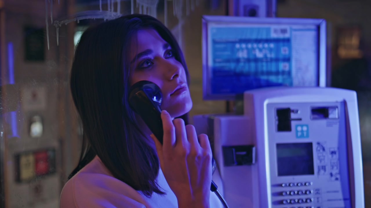 Chica morena levanta el auricular de un teléfono público en una cabina de neón en la calle. Primer plano de llamada nocturna.