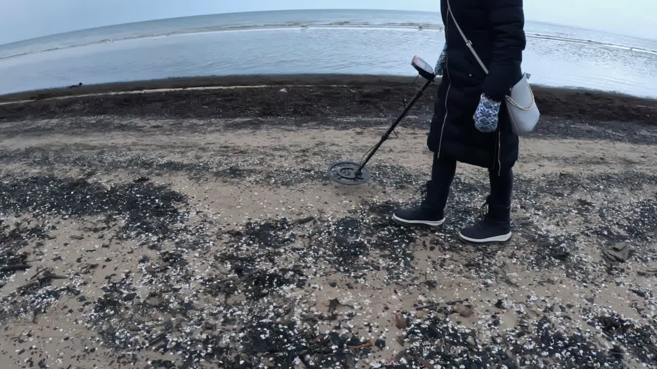 Pessoa escaneando uma costa coberta de conchas com um detector de metais em um dia frio no litoral, movendo lentamente o dispositivo sobre a areia perto da linha d'água