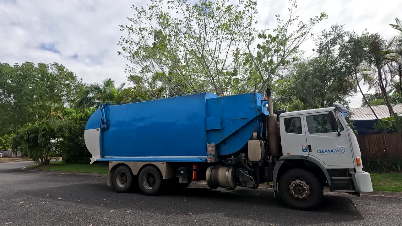Camion della spazzatura blu che passa davanti alle case in una tranquilla strada di periferia, luce del giorno, telecamera statica, cielo nuvoloso