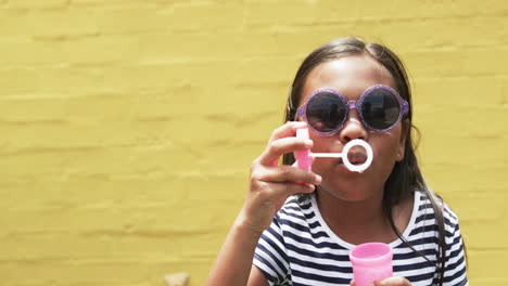 In-school,-a-young-biracial-girl-is-blowing-bubbles,-yellow-background-with-copy-space