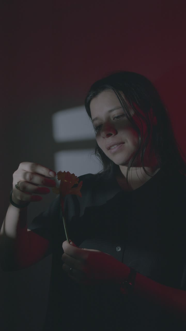 In a dimly lit room, a Hispanic girl delicately picks petals from a flower, playing 'He Loves Me, He Loves Me Not'. The soft lighting casts a warm glow, captured in vertical video format