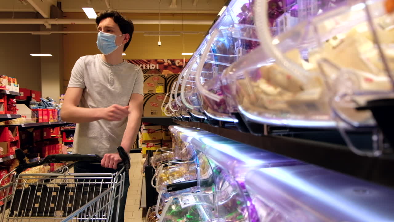CHISINAU, MOLDOVA - JUNE 5, 2020: Young man with a protective medical mask walking in a supermarket and looking in sweets dispensers. Corona Virus idea