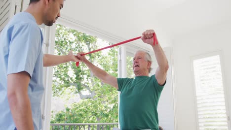 Male-physiotherapist-examining-retired-senior-man-exercising-with-resistance-band-at-retirement-home