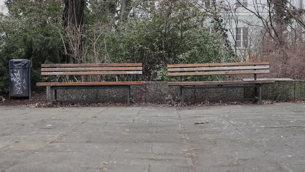 Berlin city park with two benches and a trash can on a crooked pavement under cloudy daylight