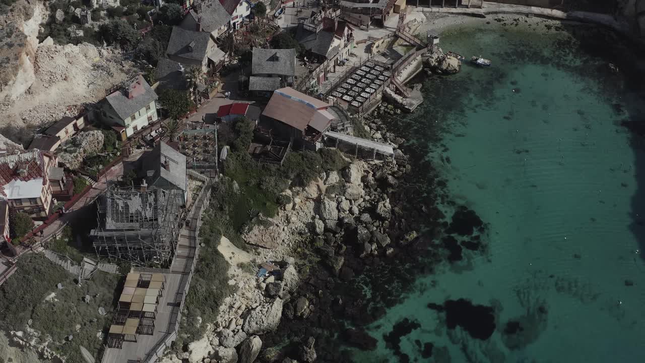 A drone flies straight alongside Popeye Village, then gradually tilts the camera downward, revealing vibrant wooden houses, Anchor Bay’s turquoise waters, and Malta’s picturesque coastline