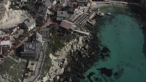 a drone flies straight alongside popeye village, then gradually tilts the camera downward, revealing vibrant wooden houses, anchor bay’s turquoise waters, and malta’s picturesque coastline