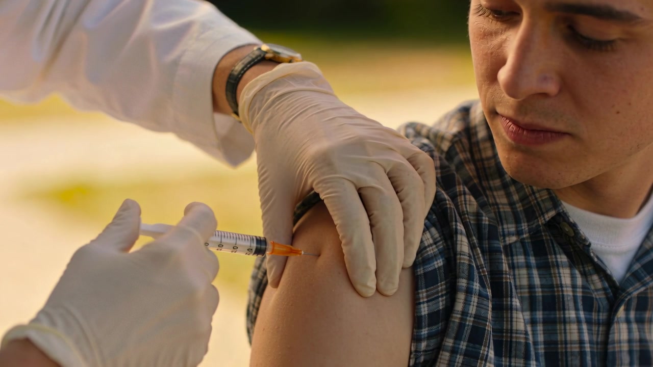 Bringing syringe, gloved nurse steadying teen's plaid sleeve outdoors, giving vaccine in upper arm