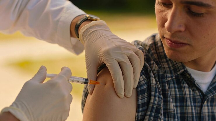 Bringing syringe, gloved nurse steadying teen's plaid sleeve outdoors, giving vaccine in upper arm