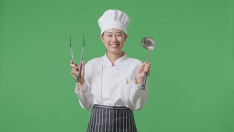 Asian Woman Chef Smiling And Showing Tongs And Ladle To Camera While Standing In The Green Screen Background Studio