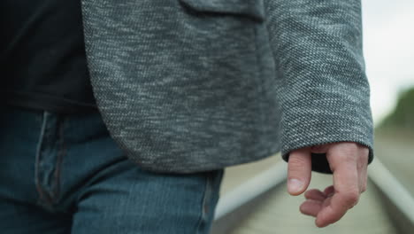 una vista de cerca de la mano de un hombre y la manga de una chaqueta de traje gris, caminando por una vía ferroviaria, el entorno está borroso, su traje incluye un blazer gris y pantalones vaqueros azules