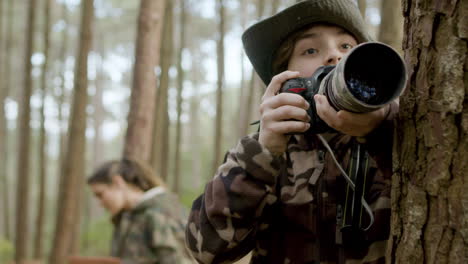 niño pequeño activo tomando fotos en el parque natural usando equipo de fotos profesional, luego mostrándole una foto a mamá sentada en el fondo y tomando notas en un libro