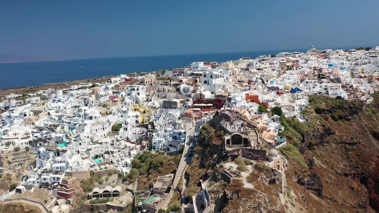 Bela tomada ampla de Oia, Santorini, apresentando casas brancas icônicas, igrejas de cúpulas azuis e dramáticas falésias vulcânicas vermelhas que descem em direção ao azul profundo do Mar Mediterrâneo em um dia claro.