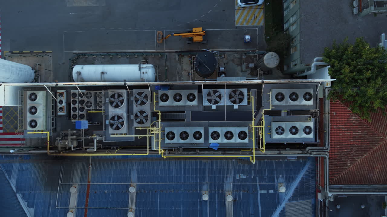 Top-down view of industrial rooftop HVAC system with multiple condenser fan units, pressure vessels, yellow piping network, corrugated metal roof panels and adjacent tiled rooftop