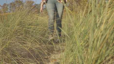 Handheld-shot-of-young-woman-walking-down-a-sand-path-toward-a-beach-at-sunset