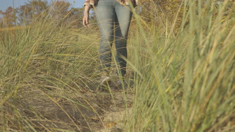 handheld shot of young woman walking down a sand path toward a beach at sunset