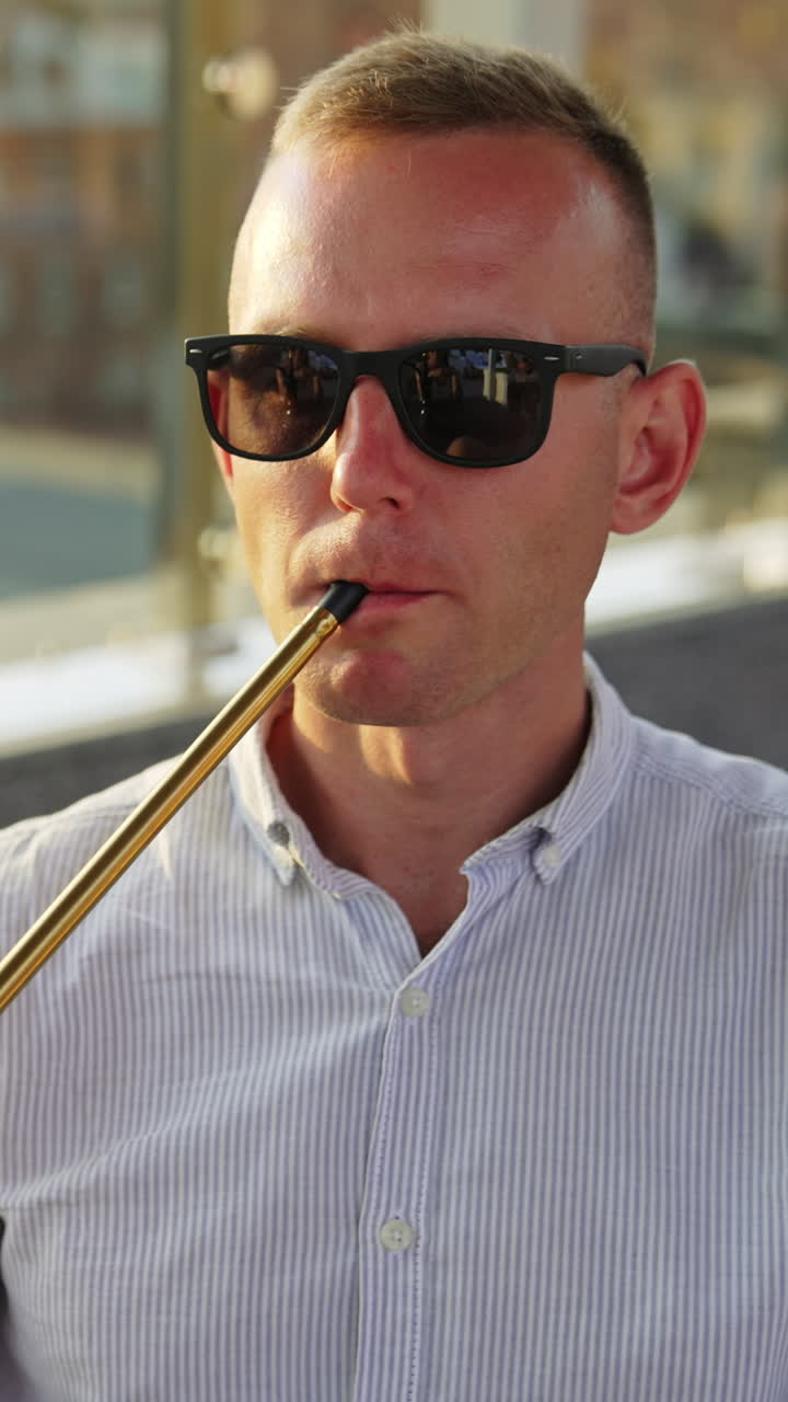 Mid aged man wearing sunglasses and shirt sits outdoors. Male resting and smoking hookah producing a cloud of thick white smoke. Vertical video