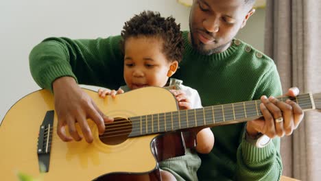 Vista-Frontal-De-Un-Joven-Padre-Negro-Y-Un-Hijo-Pequeño-Tocando-La-Guitarra-En-La-Sala-De-Estar-De-Una-Cómoda-Casa-4k