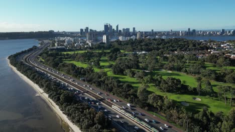 Highway-traffic-and-passing-passenger-trains-towards-the-city-beside-golf-course,-dynamic-aerial