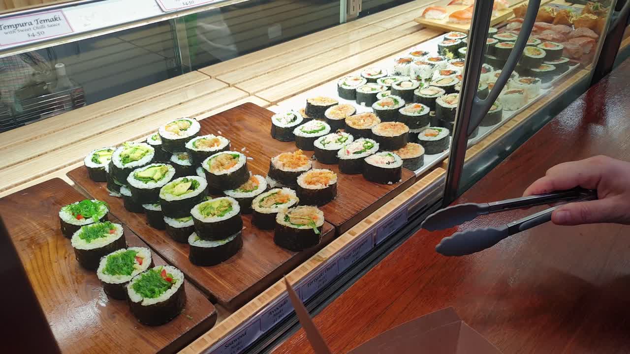 una persona seleccionando y recogiendo sus deliciosos rollos de sushi favoritos recién hechos con pinzas de un carrito local de sushi para llevar