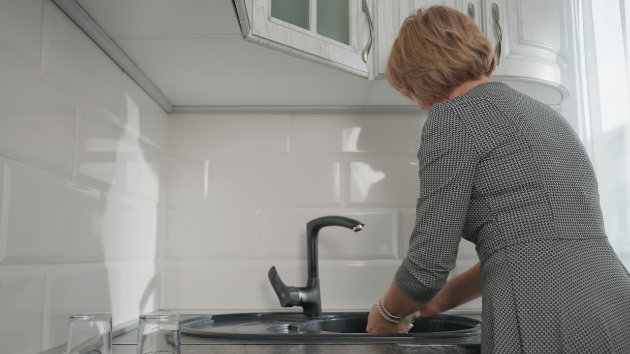 House wife drops one glass cup on kitchen counter and picks another to begin washing at sink in clean modern kitchen showing multitasking, cleanliness routine, and domestic responsibility