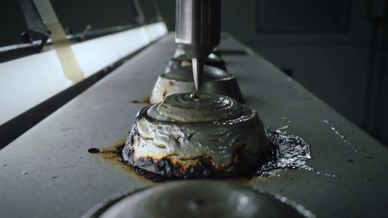 Lowering robotic nozzle depositing viscous paste onto metal dome at workbench, forming cap