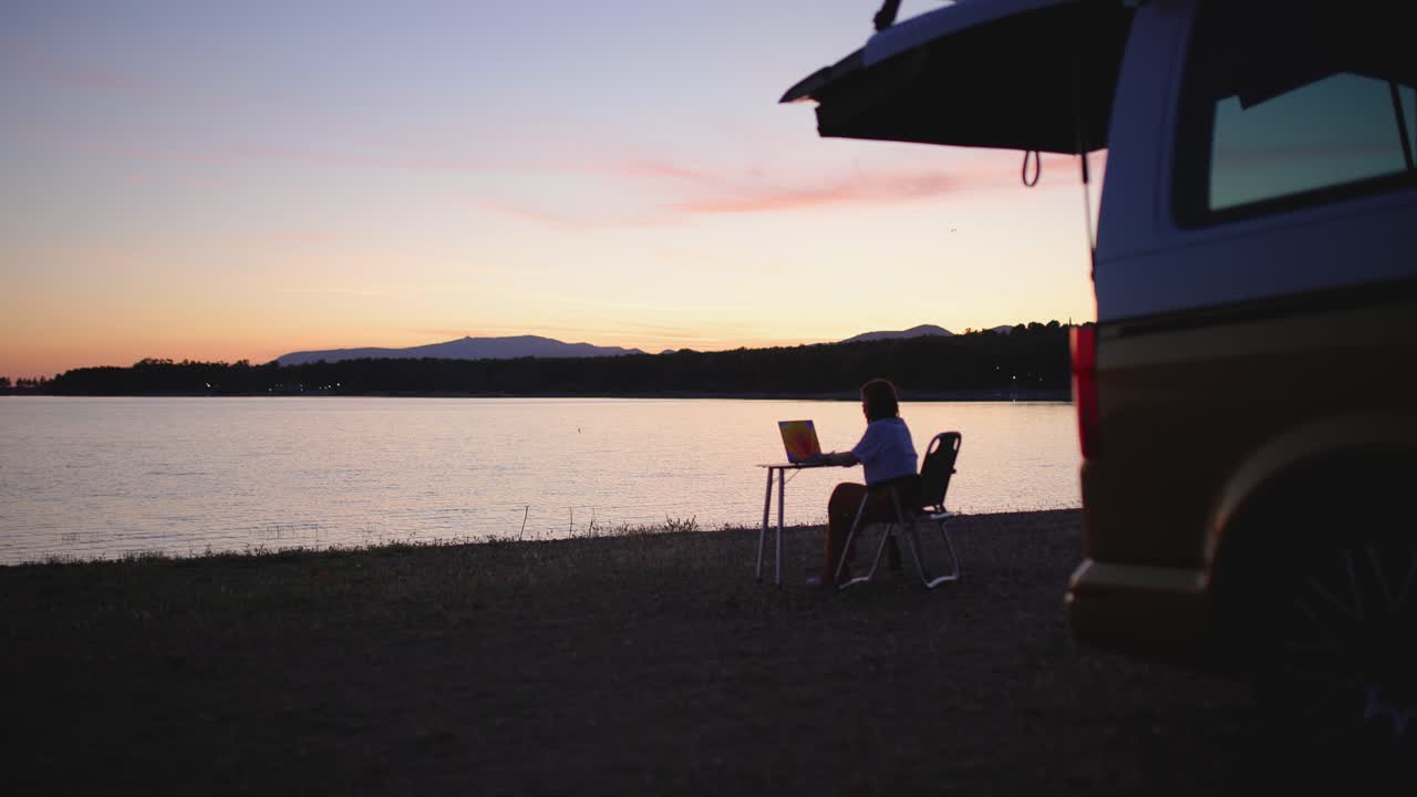 Donna che lavora al laptop in riva al lago al tramonto con un camper