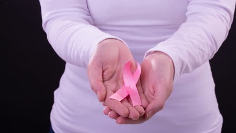 Video-of-midsection-of-caucasian-woman-holding-pink-cancer-awareness-ribbon,-with-black-background