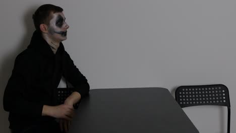 joven caucásico con maquillaje de halloween de payaso oscuro sentado solo y mirando a la pared