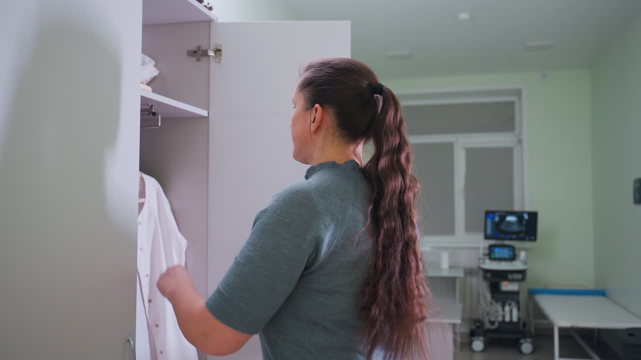 Female medical consultant walks toward locker takes out work cloth closes locker prepares for duty inside clean medical room equipped with desktop computer, ultrasound machine, and examination bed