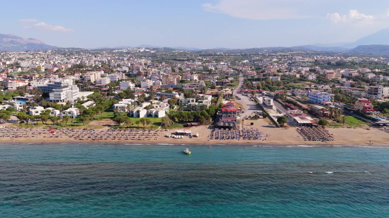 Ammoudara, Yunanistan'da, büyük kavisli bir sahil otelinin ve bir kasabanın altında şemsiyeli kumlu bir plajın havadan drone görüntüsü