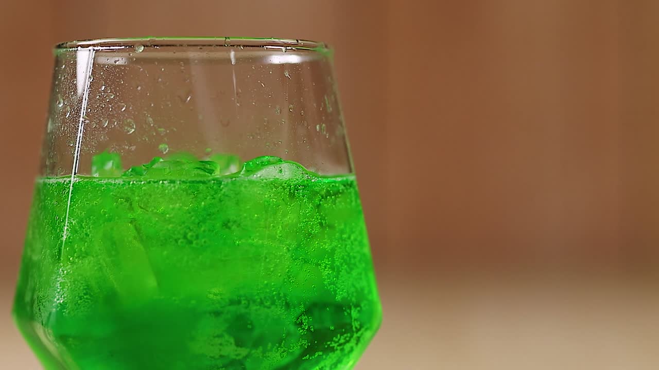 A bright green beverage with ice cubes in a uniquely shaped glass against a blurred background.